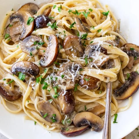 A fork twirls linguine coated in a light cream sauce with tender mushrooms and shaved Parmesan.  