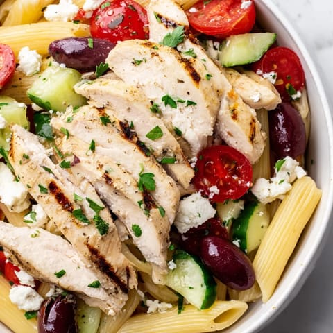 Bright and colorful Greek Chicken Pasta Bowl with lemony grilled chicken, cherry tomatoes, cucumbers, Kalamata olives, and crumbled feta on a white marble surface.