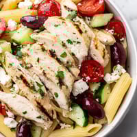 Bright and colorful Greek Chicken Pasta Bowl with lemony grilled chicken, cherry tomatoes, cucumbers, Kalamata olives, and crumbled feta on a white marble surface.